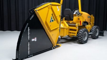 1779234033_63606_en_42fd5_65724_vermeer-mtr726-microtrencherjpg - Machines Wiki Yellow Vermeer snow plow attachment mounted on a wheeled tractor indoors, blade raised against a dark backdrop.
