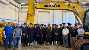 Group of graduates in black robes and mortarboards posing in a factory beside a large yellow Komatsu excavator.