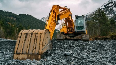 Yellow excavator on a rubble-strewn construction site with a large digging bucket, mountains in the background.