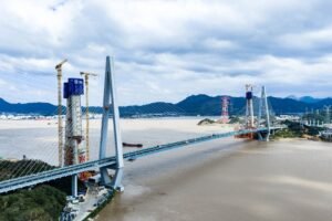 Suspension/cable-stayed bridge under construction over a wide river, with cranes and tall pylons against a cloudy sky and distant mountains.