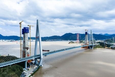 Suspension/cable-stayed bridge under construction over a wide river, with cranes and tall pylons against a cloudy sky and distant mountains.