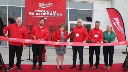 Six people in red Milwaukee shirts cut a red ribbon at a grand-opening event on a red carpet, with a large red banner behind them that reads 'Proud to be in Georgina' and Milwaukee flags nearby.