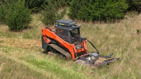 1779493483_63695_en_c379d_65826_loftness-kwik-cut-brush-cutterjpg - Machines Wiki Orange Kubota compact track loader with a front-mounted mower attachment cutting grass on a hillside field in woodland edge landscape.