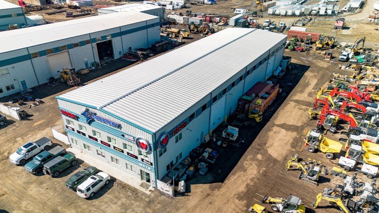 1779742081_63644_en_f2055_65769_jt-equipment-outdoor-shopjpg - Machines Wiki Aerial view of a long blue metal warehouse with a white roof, surrounded by vehicles and construction equipment in an outdoor yard.