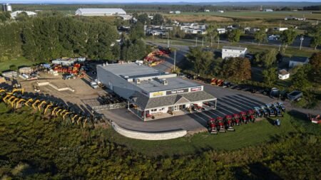Aerial view of an agricultural equipment dealership: a gray showroom building with outdoor displays of red tractors and yellow machines, surrounded by a parking lot, trees, and fields.