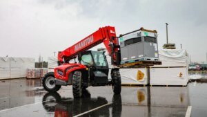 1779838272_63613_en_7ae06_65731_manitou-mta-842jpg - Machines Wiki Red Manitou telehandler lifting a large gray equipment crate on a pallet in a wet outdoor yard with stacked wrapped pallets nearby.
