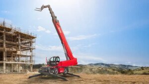 1779841903_63609_en_59bc3_65727_magni-rth-17167-telehandlerjpg - Machines Wiki Red mobile crane with an extended boom and stabilizers deployed, positioned beside a partially built concrete structure on a dirt site.