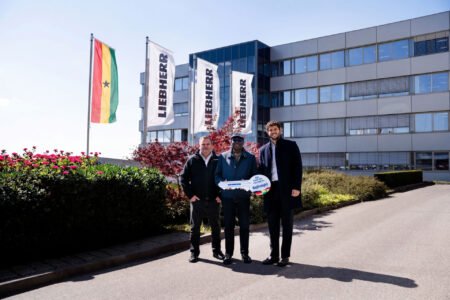 Three men stand outdoors in front of a modern office building with flagpoles and white flags; one holds a large ceremonial key crane.