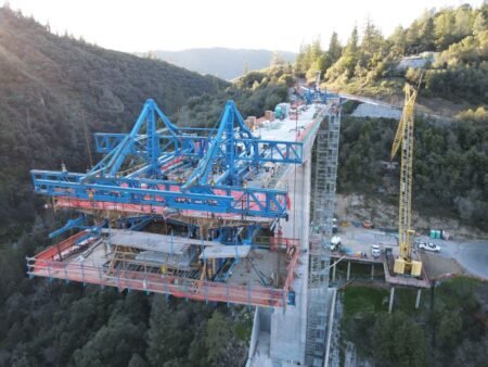 Aerial view of a large blue steel bridge under construction over a forested valley, with scaffolding and a yellow crane nearby.