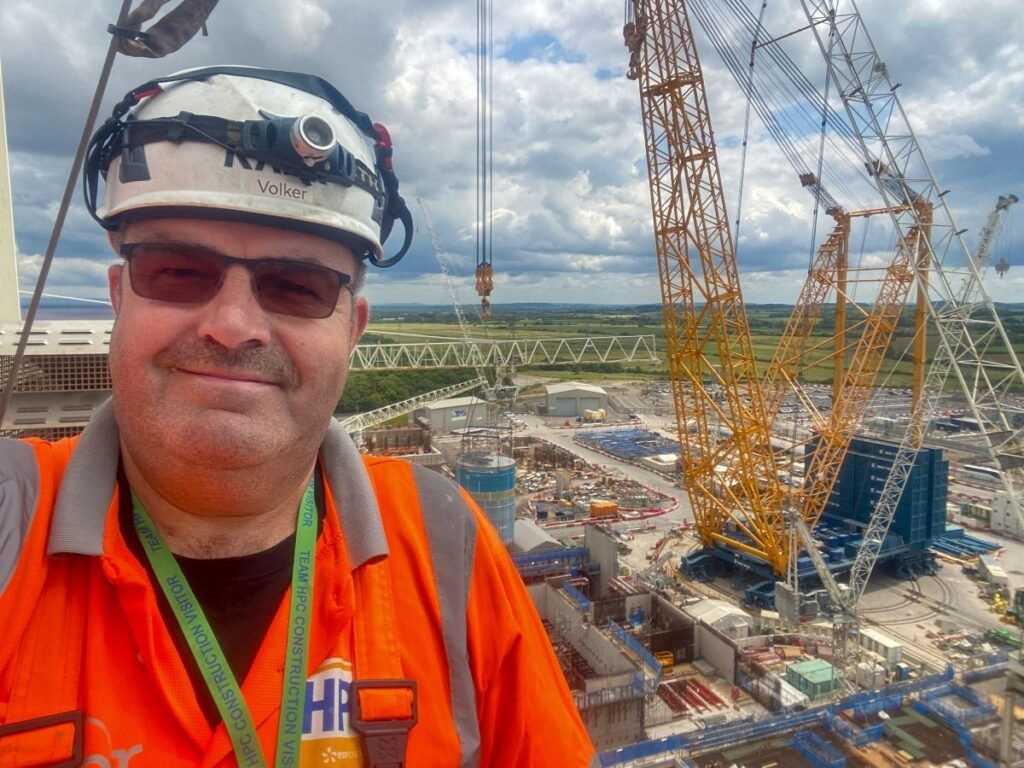 Construction worker in an orange safety vest and helmet taking a selfie on a crane platform overlooking a large construction site with a yellow lattice crane in the background.