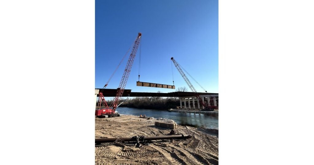 Two red cranes lift a large steel beam to place on a river bridge under construction, with blue sky overhead.