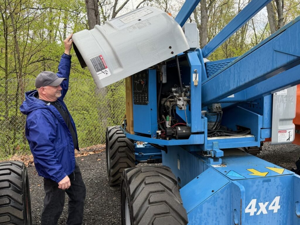 Man wearing a blue jacket and cap opening the engine compartment of a blue 4x4 aerial lift outdoors by a fence and trees.
