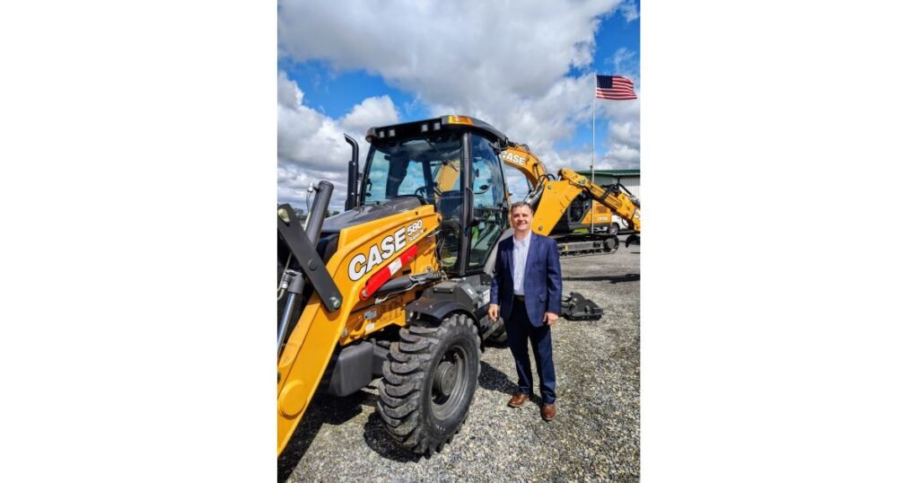 905x0_s3-71318-N-MEDICO-MUNCY-1jpg - Machines Wiki Man in a blue suit posing next to a yellow CASE backhoe loader on a gravel lot with a blue sky and clouds behind him.