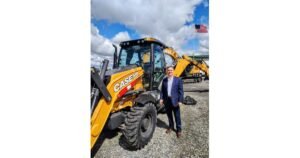 Man in a blue suit posing next to a yellow CASE backhoe loader on a gravel lot with a blue sky and clouds behind him.