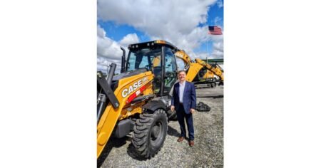 Man in a blue suit posing next to a yellow CASE backhoe loader on a gravel lot with a blue sky and clouds behind him.