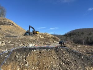 905x0_s3-71319-N-64_26-CM-1_jpegjpg - Machines Wiki Construction site on a rocky hillside with a blue pipe line, orange cones, and heavy machinery (excavator and dump truck) working on grading a road.