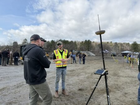 905x0_s3-71320-NE-UC_F-DEMO-DAYS-26-10_JPEGjpg - Machines Wiki Man in a high-visibility safety vest speaks with a colleague at an outdoor construction site, with survey equipment on a tripod in the foreground.