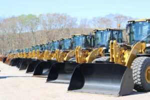 Row of yellow front-end loaders with large black buckets parked on a dirt lot, trees in a clear sky background.