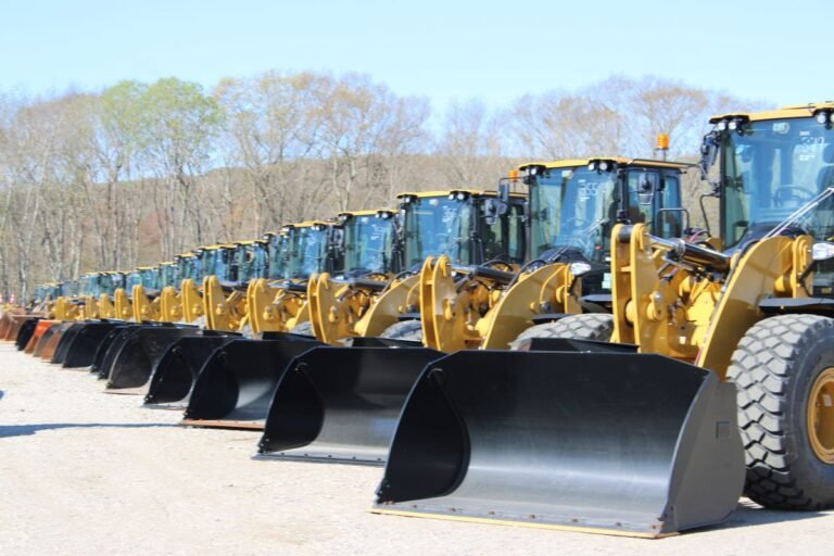 Row of yellow front-end loaders with large black buckets parked on a dirt lot, trees in a clear sky background.