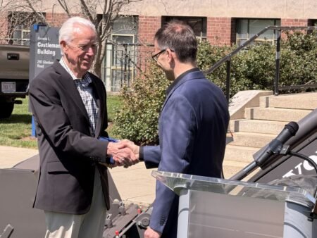 Two men in suits shake hands outside a building during a formal outdoor event, with a podium microphone nearby and a sign that reads 'Castleman Building' in the background.