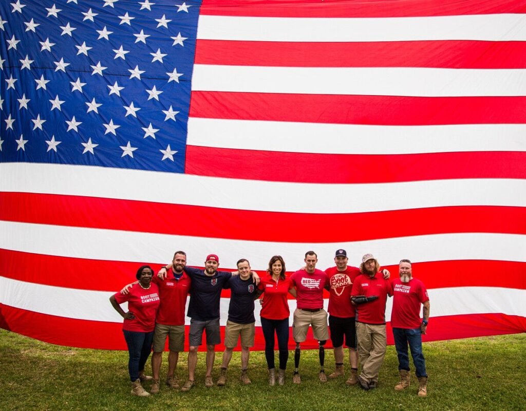 Group of people in red shirts stand arm-in-arm in front of a massive American flag outdoors, showing a patriotic team photo.