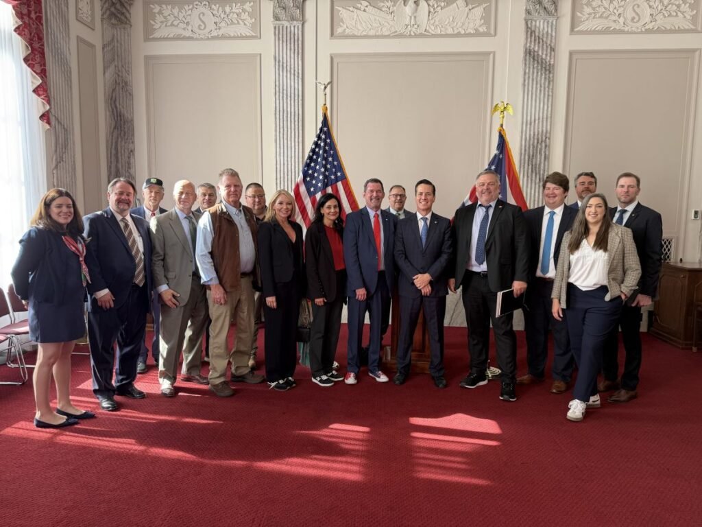 905x0_s3-71324-1000011327jpg - Machines Wiki Group of men and women in business attire posing for a photo in a formal room with two US flags.