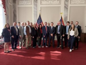 Group of men and women in business attire posing for a photo in a formal room with two US flags.
