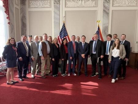 Group of men and women in business attire posing for a photo in a formal room with two US flags.