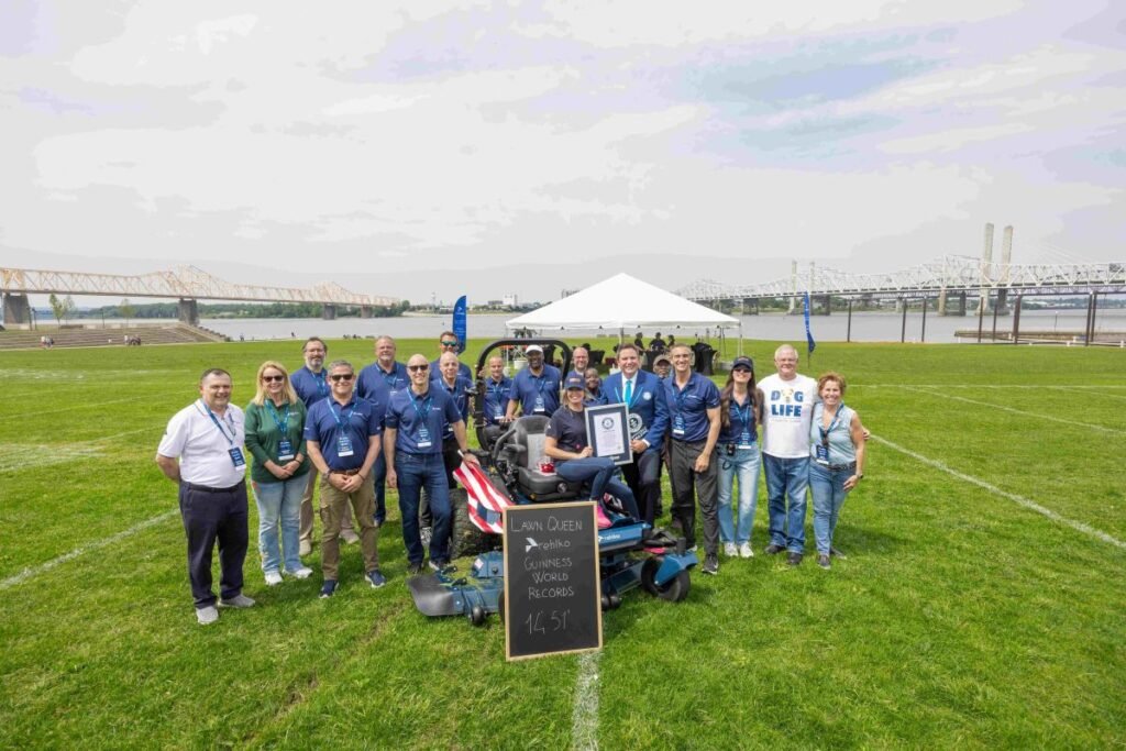 905x0_s3-71328-MNSW-TURFMUTT-RECORD-1jpg - Machines Wiki Group of people posing with a riding lawn mower and a certificate at a Guinness World Records event on a grassy field by the river.
