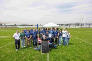 Group of people posing with a riding lawn mower and a certificate at a Guinness World Records event on a grassy field by the river.