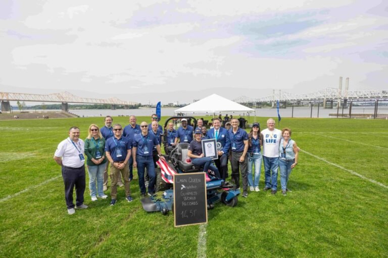 Group of people posing with a riding lawn mower and a certificate at a Guinness World Records event on a grassy field by the river.