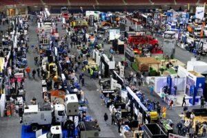 Wide view of a crowded indoor expo with numerous booths, banners and trucks, as attendees explore industrial equipment displays.
