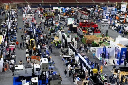 Wide view of a crowded indoor expo with numerous booths, banners and trucks, as attendees explore industrial equipment displays.
