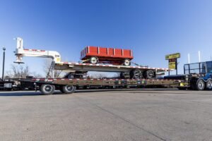 Flatbed truck trailer carrying a red wagon with a white crane on the left, in a paved lot under a blue sky.