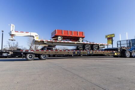 Flatbed truck trailer carrying a red wagon with a white crane on the left, in a paved lot under a blue sky.