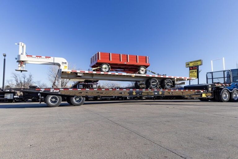 Flatbed truck trailer carrying a red wagon with a white crane on the left, in a paved lot under a blue sky.