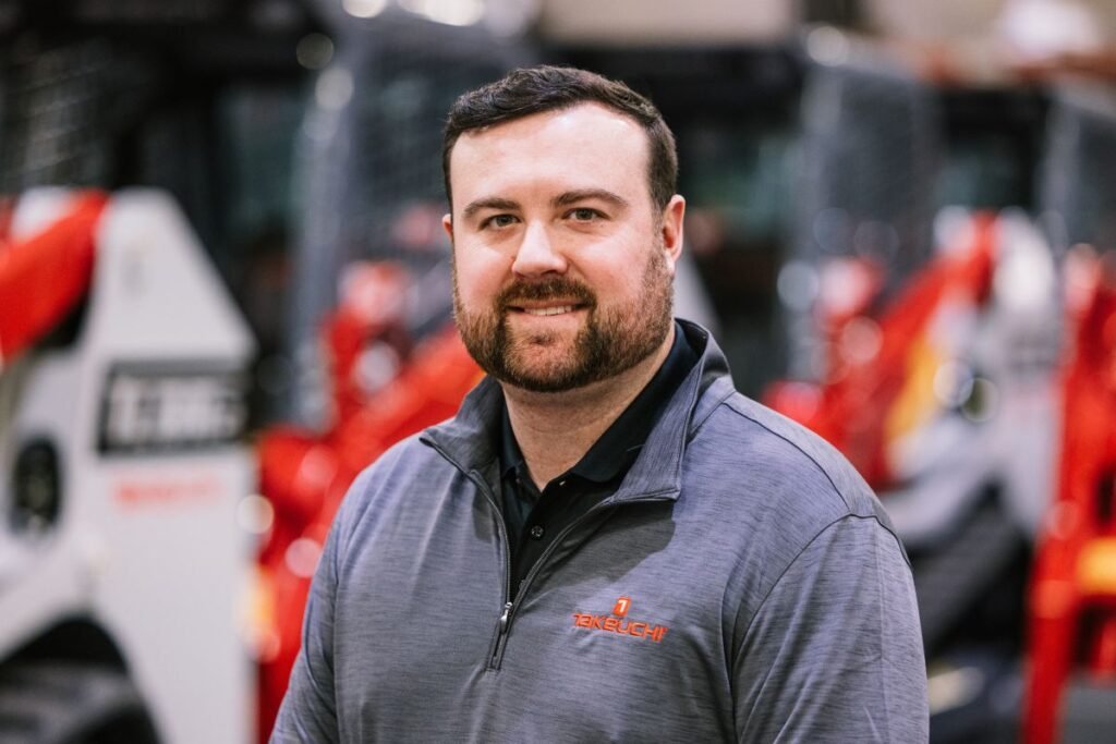 905x0_s3-71332-MNSW-TAK-NEW-REG-MGRS-1jpg - Machines Wiki Smiling man in a gray Takeuchi zip-up jacket, standing in a workshop with red machinery in the background.