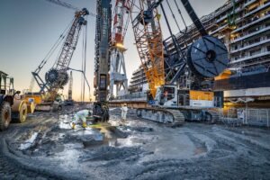 Massive drilling crane at a dockyard construction site; workers in protective gear stand in muddy ground with a ship and tall cranes overhead.