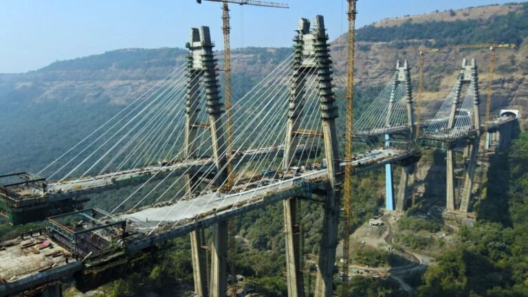 905x0_s3-71334-MNSW-CQ-POTAIN-MUMBAI-1jpg - Machines Wiki Under-construction cable-stayed bridge with tall pylons and many cables over a valley. Cranes and construction equipment around the site.