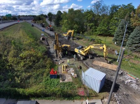 Construction site beside a road with several yellow excavators, workers, and dirt, preparing a trench area near a green wooded area
