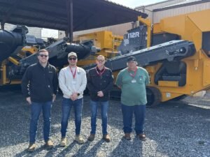 905x0_s3-71336-M-OH-STONE-ANA_JPGjpg - Machines Wiki Four workers posing in front of a large yellow industrial machine outdoors on a gravel lot.