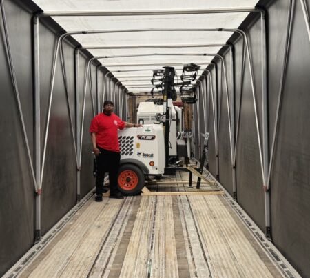 Man in a red shirt stands beside a Bobcat PL65 skid-steer loader inside an empty loading trailer with metal walls and overhead lighting.
