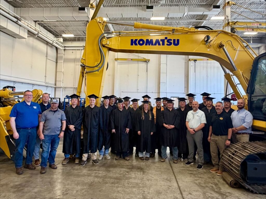 905x0_s3-71341-M-KOM-NDSCS-GRADS-1_jpegjpg - Machines Wiki Group of graduates in black gowns and mortarboards posing with instructors inside a workshop next to a large Komatsu excavator.