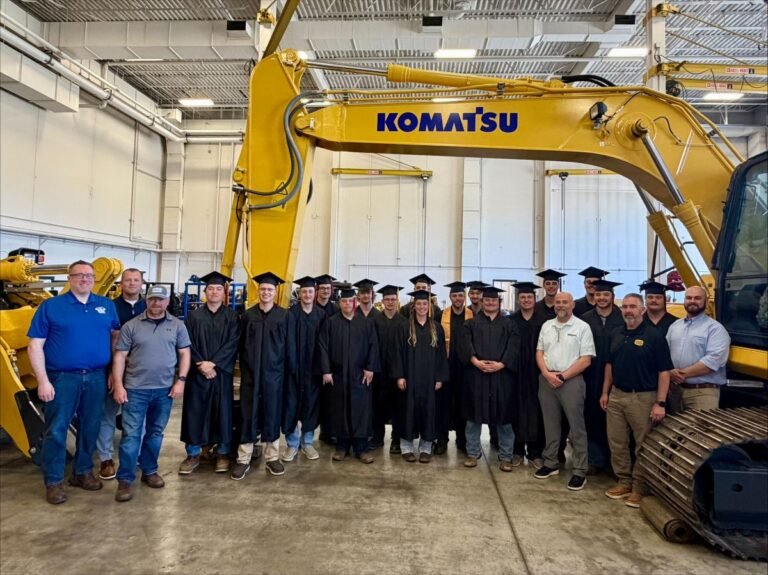 Group of graduates in black gowns and mortarboards posing with instructors inside a workshop next to a large Komatsu excavator.