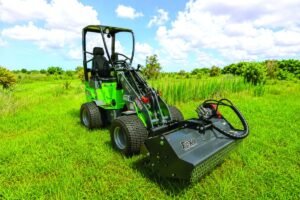 905x0_s3-71343-MNSW-ATT-FOXX-LINEUP-1jpg - Machines Wiki Green compact skid-steer loader with a large front attachment in a grassy field under a bright blue sky, ready for work.