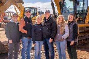 Six construction workers stand together in front of yellow excavators on a dirt job site, smiling at the camera.