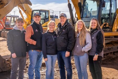 Six construction workers stand together in front of yellow excavators on a dirt job site, smiling at the camera.
