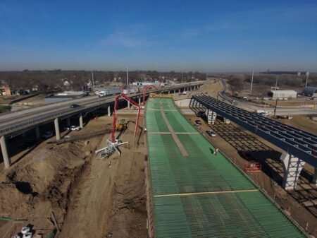 905x0_s3-71350-M-066_26-CR-1_JPGjpg - Machines Wiki Aerial view of a bridge construction site with green deck formwork, a red concrete pump, and nearby elevated highways under a clear blue sky.