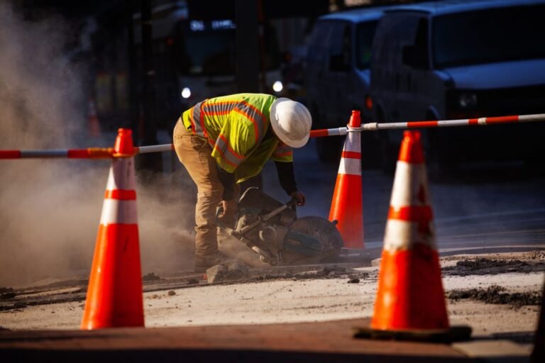 Getting Every Cent Out of Projects : CEG Construction worker in a neon safety vest and hard hat operates a cut-off saw on a dusty road, orange traffic cones surrounding the work area.
