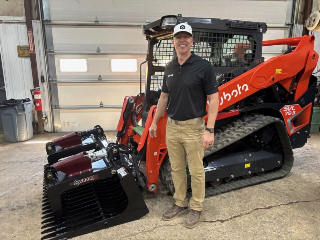 905x0_s3-71357-M-LANO-CAD-26-1jpg - Machines Wiki Man in a black polo and khaki pants standing beside a red skid-steer loader with a large black grapple attachment in a workshop.